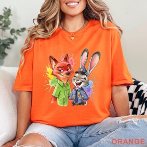 May include: Orange t-shirt featuring a cartoon fox and rabbit with colorful paint splatter accents. The fox wears a green shirt, and the rabbit is in a police uniform. The word "ORANGE" is printed at the bottom of the shirt.