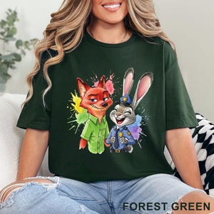 May include: Forest green t-shirt featuring a colorful graphic of a fox and a rabbit. The fox is wearing a green shirt, and the rabbit is in a police uniform. The shirt has the words "FOREST GREEN" printed at the bottom.