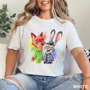 May include: White t-shirt featuring a colorful illustration of Nick Wilde the fox and Judy Hopps the rabbit from Zootopia. Nick is wearing a green shirt, and Judy is in a police uniform. The word "WHITE" is printed at the bottom.