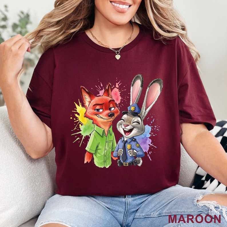 May include: Maroon t-shirt featuring a colorful graphic of Nick Wilde the fox and Judy Hopps the rabbit from Zootopia. Nick is wearing a green shirt, and Judy is in a police uniform. The word "MAROON" is printed at the bottom.
