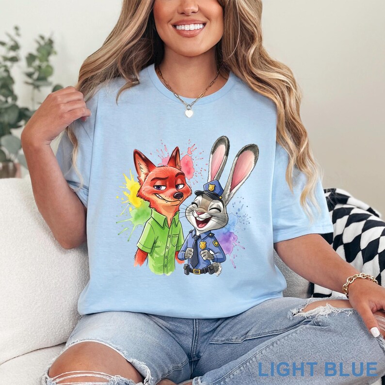 May include: Light blue t-shirt featuring a colorful graphic of Nick Wilde and Judy Hopps from Zootopia. Nick is wearing a green shirt, and Judy is in a police uniform. The text "LIGHT BLUE" is at the bottom.