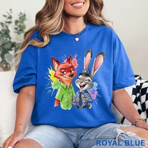 May include: Royal blue t-shirt featuring a colorful graphic of Nick Wilde the fox and Judy Hopps the rabbit from Zootopia. The shirt has a crew neck and short sleeves. The text "ROYAL BLUE" is printed at the bottom.