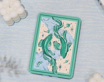 May include: A rectangular, mint green coaster featuring a whimsical illustration of two turquoise fish swimming amidst clouds, stars, and planets. The background is a soft beige with light blue clouds. The coaster has a mint green border.