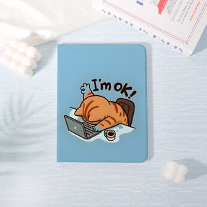 May include: A light blue notebook with a cartoon illustration of a cat working on a laptop. The cat is orange and brown, with the text "I'm ok!" above it. A small coffee cup is next to the laptop.