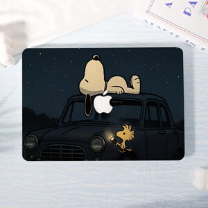 May include: A laptop case featuring a cartoon illustration of Snoopy lying on top of a black car under a starry night sky. Woodstock is holding a flashlight. The Apple logo is visible on the car.