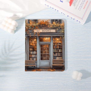 Peut inclure: Un carnet rectangulaire présentant une illustration imprimée de la devanture d'une librairie avec une enseigne indiquant BOOKS. La façade est peinte dans des tons bleu sourd, ornée de feuillage d'automne et flanquée de deux bancs en bois.