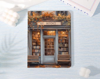May include: A rectangular notebook featuring a printed illustration of a bookstore storefront with a sign that reads BOOKS. The facade is painted in muted blue tones, adorned with autumn foliage, and flanked by two wooden benches on either side.