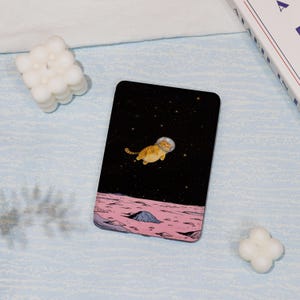 May include: A digital illustration of a cat wearing a space helmet floating in space above a pink and gray planet. The background is a black starry sky.