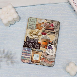 May include: A phone case with a collage design featuring books, coffee cups, pumpkins, and a ghost reading a book. The text includes "I have lived a thousand lives" and "I read past my bedtime". The case has a beige background.