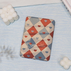 May include: A rectangular, patterned item with a red, blue, and cream diamond design, featuring heart and flower motifs. The word "HAPPY" is printed on the bottom with a smiling face. The item is on a light blue surface.