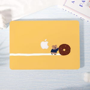 May include: Yellow laptop case with a white Apple logo and a cartoon bear pushing a chocolate donut. A white stripe runs across the bottom of the case. The case is on a light blue surface.