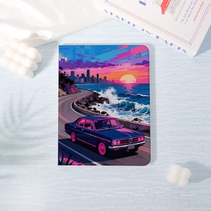 Könnte beinhalten: Ein Notizbuch mit einer farbenfrohen Illustration eines Oldtimers auf einer Küstenstraße bei Sonnenuntergang. Das Kunstwerk hat einen Retro-Stil mit Blau-, Rosa- und Orangetönen, die ein Gefühl von Nostalgie und Abenteuer erzeugen. Ein stilvolles Accessoire.