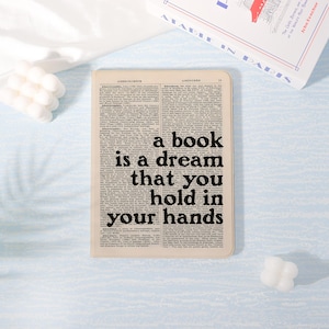 May include: A notebook featuring a vintage dictionary page design. The cover displays the quote "a book is a dream that you hold in your hands" in black. A book and two white decorative candles are also in the image.
