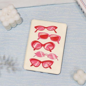 May include: A cream-coloured rectangular item displaying five pairs of pink and red sunglasses. The sunglasses are in various styles, including cat-eye, square, and oval shapes. The background is a light blue surface.