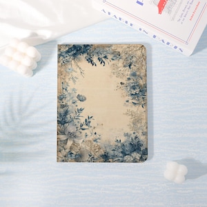 May include: A rectangular journal with a floral design. The cover features a cream-colored background with blue and gray floral illustrations. The journal is surrounded by a white candle and a book titled "Paris".