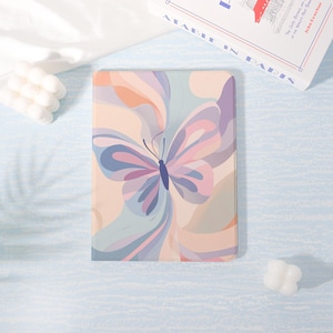 May include: A pastel-colored tablet case featuring a butterfly design. The butterfly has blue, purple, and pink wings with a dark blue body. The background has flowing abstract shapes in peach, lavender, and blue.
