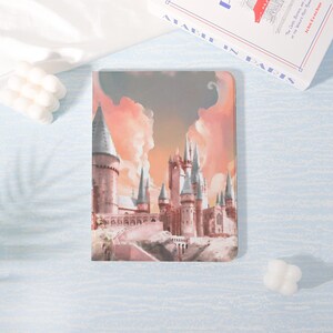 May include: A rectangular case with a castle illustration under a pink and orange sky. The castle has multiple towers and spires. The case is on a light blue surface with a white candle and a book in the background.