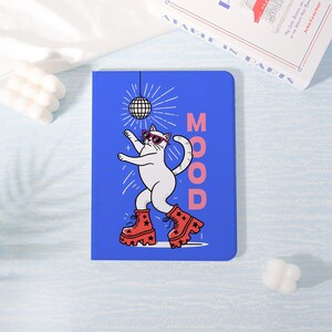 May include: A blue notebook featuring a white cat in sunglasses and red boots dancing under a disco ball. The word "MOOD" is printed vertically in pink. A book and a white decorative object are in the background.