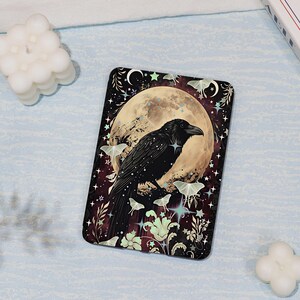 Peut inclure: Une housse de liseuse rectangulaire illustrée d'un corbeau noir perché devant une grande pleine lune. Le motif comprend des papillons de nuit vert pâle, des étoiles et des fleurs sur un fond violet foncé et noir avec des croissants de lune.