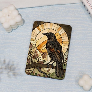 May include: A rectangular e-reader cover featuring a stained glass style illustration of a black crow perched on a flowering branch. The background displays a circular golden sun motif with geometric lines, set against a light blue textured surface.