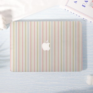 May include: A laptop with a colorful striped design. The cover features vertical stripes in pastel pink, blue, and green on a cream background. The Apple logo is visible in the center. A book is visible in the background.
