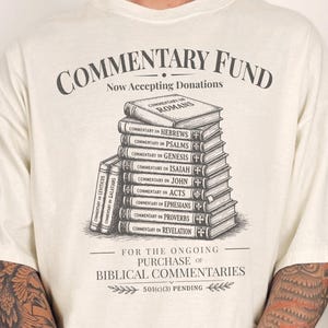 Może przedstawiać: Kremowa koszulka z grafiką ułożonych w stos książek z tytułami komentarzy biblijnych. Koszulka zawiera tekst "COMMENTARY FUND" i "Now Accepting Donations" w stylu vintage.