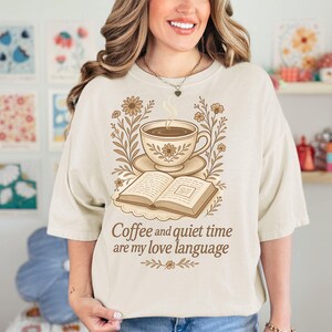May include: Cream-colored t-shirt featuring a coffee cup, saucer, and open book illustration with floral accents. The text "Coffee and quiet time are my love language" is printed below the design. The shirt has short sleeves.