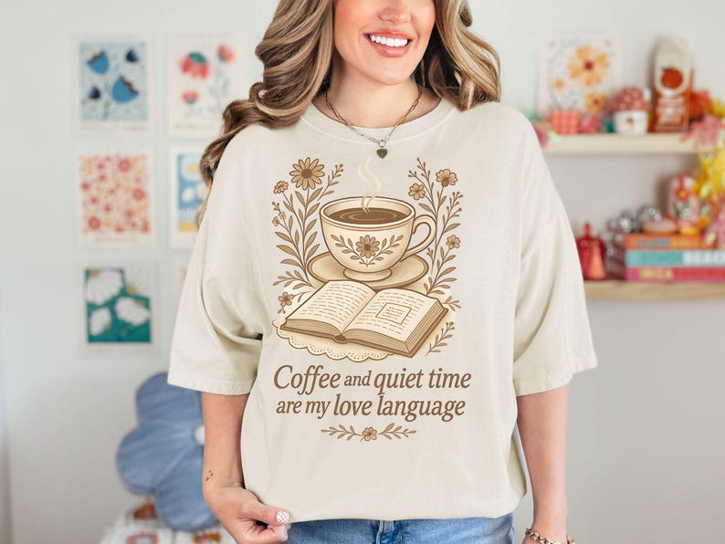 May include: Cream-colored t-shirt featuring a coffee cup, saucer, and open book illustration with floral accents. The text "Coffee and quiet time are my love language" is printed below the design. The shirt has short sleeves.