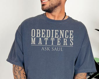 May include: A navy blue t-shirt with the text "OBEDIENCE MATTERS ASK SAUL" in a white, bold font. The shirt has short sleeves and a crew neck. The image shows the upper body of a person wearing the shirt.