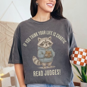 May include: Dark gray t-shirt featuring a cartoon raccoon holding a book, with the text "IF YOU THINK YOUR LIFE IS CHAOTIC READ JUDGES!". The raccoon wears a blue hoodie. A checkered orange and white mug is held in the hand.