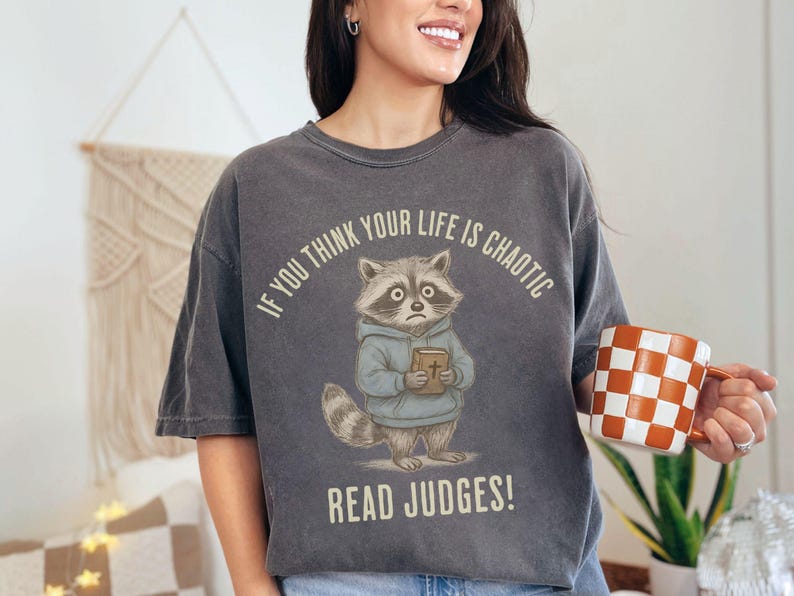 May include: Dark gray t-shirt featuring a cartoon raccoon holding a book, with the text "IF YOU THINK YOUR LIFE IS CHAOTIC READ JUDGES!". The raccoon wears a blue hoodie. A checkered orange and white mug is held in the hand.