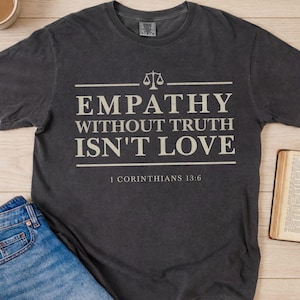 May include: Dark gray t-shirt with the text "EMPATHY WITHOUT TRUTH ISN'T LOVE" and "1 CORINTHIANS 13:6" in white. The design includes a scale icon. Blue jeans and an open book are also visible.