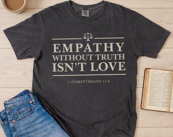 Op de afbeelding: Donkergrijs t-shirt met de witte tekst "EMPATHY WITHOUT TRUTH ISN'T LOVE" en "1 CORINTHIANS 13:6". Het ontwerp bevat een weegschaal-icoon. Een blauwe jeans en een open boek zijn ook zichtbaar.