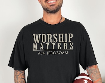 Op de afbeelding: Zwart T-shirt met de tekst "WORSHIP MATTERS ASK JEROBOAM" in lichtbeige. Het shirt is donker van kleur en heeft korte mouwen. Een voetbal wordt in de handen van de persoon gehouden.