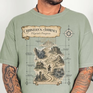 May include: A sage green t-shirt featuring a detailed map illustration titled Christian's Journey Pilgrim's Progress. The graphic depicts a winding path through various landmarks like the Celestial City, Doubting Castle, and Valley of the Shadow of Death.