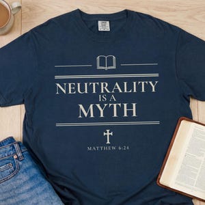 May include: Navy blue t-shirt with the text "NEUTRALITY IS A MYTH" and a cross graphic. The shirt is paired with blue jeans, an open book, and a mug of coffee. The text "Matthew 6:24" is also printed on the shirt.