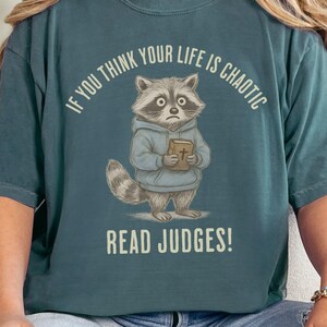 May include: Teal-colored t-shirt featuring a cartoon raccoon wearing a blue hoodie and holding a book. The text "IF YOU THINK YOUR LIFE IS CHAOTIC READ JUDGES!" is printed above and below the raccoon illustration. The shirt has short sleeves.