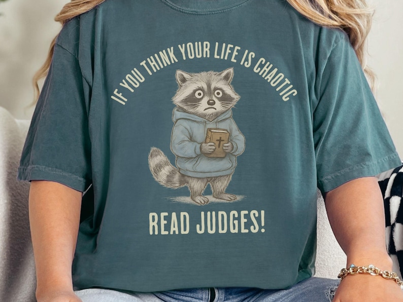 May include: Teal-colored t-shirt featuring a cartoon raccoon wearing a blue hoodie and holding a book. The text "IF YOU THINK YOUR LIFE IS CHAOTIC READ JUDGES!" is printed above and below the raccoon illustration. The shirt has short sleeves.