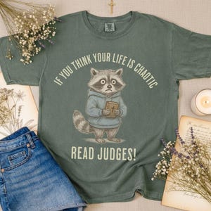 May include: Sage green t-shirt featuring a raccoon wearing a hoodie and holding a book with a cross. The text reads "IF YOU THINK YOUR LIFE IS CHAOTIC READ JUDGES!" with a distressed design. The shirt is paired with denim shorts.