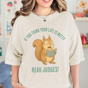 May include: Cream-colored t-shirt featuring a cartoon squirrel reading a book with the text "IF YOU THINK YOUR LIFE IS NUTTY READ JUDGES!". The squirrel is brown with a surprised expression.