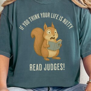 May include: Teal-colored t-shirt featuring a cartoon squirrel reading a book with the text "IF YOU THINK YOUR LIFE IS NUTTY READ JUDGES!". The squirrel is brown with a surprised expression. The text is in white.