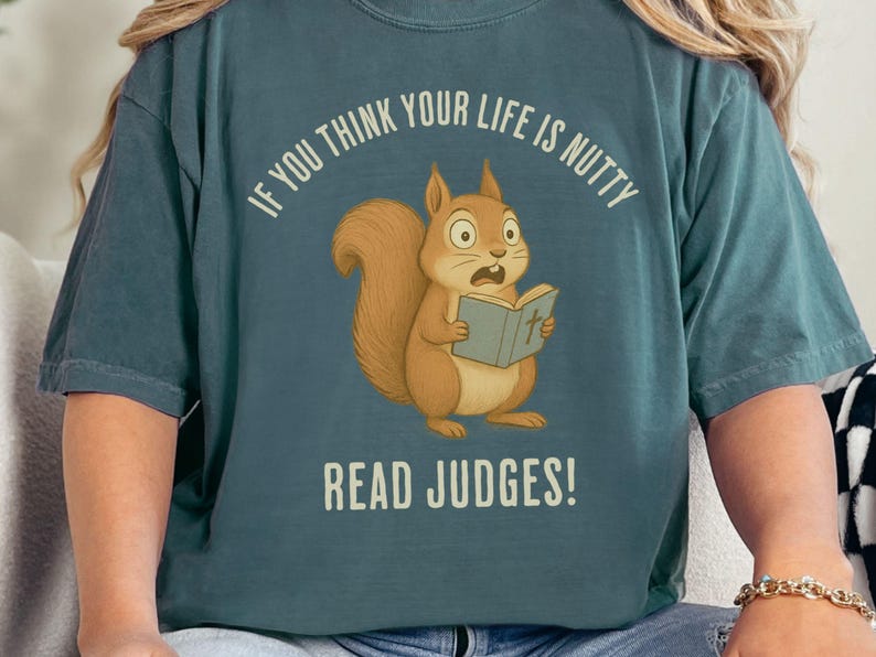 May include: Teal-colored t-shirt featuring a cartoon squirrel reading a book with the text "IF YOU THINK YOUR LIFE IS NUTTY READ JUDGES!". The squirrel is brown with a surprised expression. The text is in white.