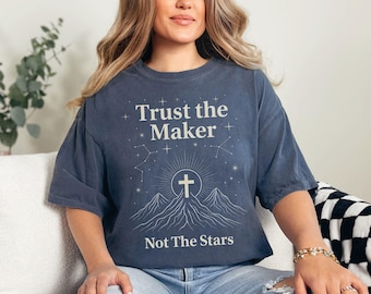 Op de afbeelding: Donkerblauw T-shirt met de tekst "Trust the Maker, Not The Stars". Het shirt heeft een afbeelding van een kruis, bergen en sterrenbeelden. Het shirt is gemaakt van een zacht materiaal.