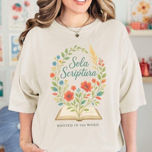 Puede incluir: Camiseta color crema con un diseño de corona floral y la inscripción "Sola Scriptura". El diseño incluye elementos rojos, naranjas, azules y verdes, con la ilustración de un libro abierto y el texto "Rooted in the Word" debajo.