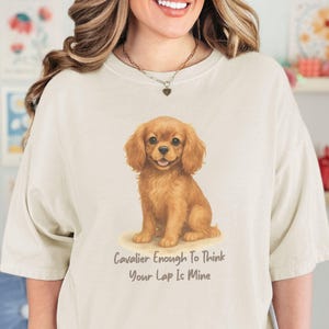Pode incluir: Camiseta creme com uma ilustração em aquarela de um filhote de Cavalier King Charles Spaniel. O texto "Cavalier Enough To Think Your Lap Is Mine" está impresso abaixo. A camiseta tem um corte folgado.