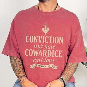 Op de afbeelding: Rood t-shirt met de tekst "CONVICTION isn't hate COWARDICE isn't love" in lichtbeige. Het ontwerp bevat een hart doorboord door een zwaard en de tekst "Ephesians 4:15" op een banner.
