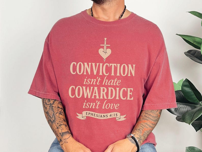 Op de afbeelding: Rood t-shirt met de tekst "CONVICTION isn't hate COWARDICE isn't love" in lichtbeige. Het ontwerp bevat een hart doorboord door een zwaard en de tekst "Ephesians 4:15" op een banner.