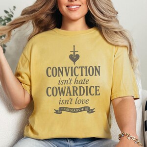 Op de afbeelding: Mosterdgeel T-shirt met de grijze tekst "CONVICTION isn't hate, COWARDICE isn't love". Het ontwerp bevat een hart doorboord door een zwaard. De tekst "Ephesians 4:15" staat onder het citaat.