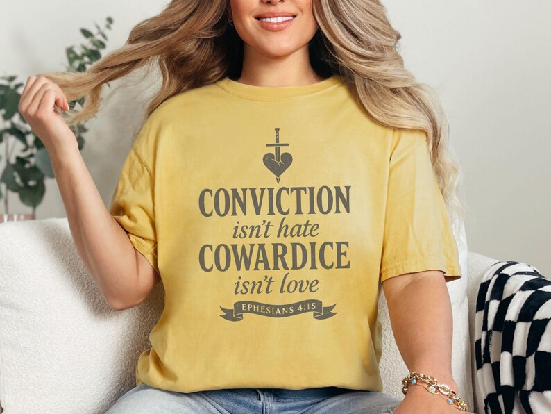 Op de afbeelding: Mosterdgeel T-shirt met de grijze tekst "CONVICTION isn't hate, COWARDICE isn't love". Het ontwerp bevat een hart doorboord door een zwaard. De tekst "Ephesians 4:15" staat onder het citaat.