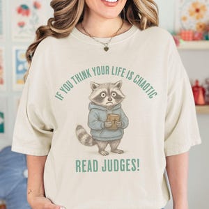 May include: Cream-colored t-shirt featuring a cartoon raccoon wearing a blue hoodie and holding a book. The text "IF YOU THINK YOUR LIFE IS CHAOTIC" arches above the raccoon, with "READ JUDGES!" below.